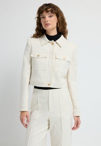 Cremefarvet cropped jakke med krave, to forlommer og guldknapdetaljer. Båret over en sort top og matchende bukser.