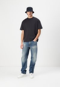Black cotton t-shirt, loose fit. Light blue distressed denim jeans. White sneakers. Navy bucket hat. Model standing against a plain background.