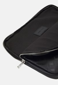 Black laptop sleeve featuring a smooth exterior with a zip closure, a fabric interior, and a logo patch. Textured pattern on the lining.