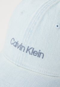 Šviesiai mėlynas džinsinis kepurė su lenktu kraštu, su siūtais aklais ir tamsiai mėlyna siuvinėta "Calvin Klein" užrašu.