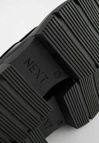 Schwarze Gummisohle mit tiefen Profilen und Rillen, geprägt mit "NEXT" und Größenangaben. Das Material wirkt langlebig mit strukturierter Oberfläche.