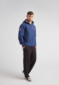 Veste imperméable marine avec doublure intérieure grise, zip sur le devant, deux poches latérales et écusson logo sur la manche ; assortie avec un pantalon large noir et des baskets.