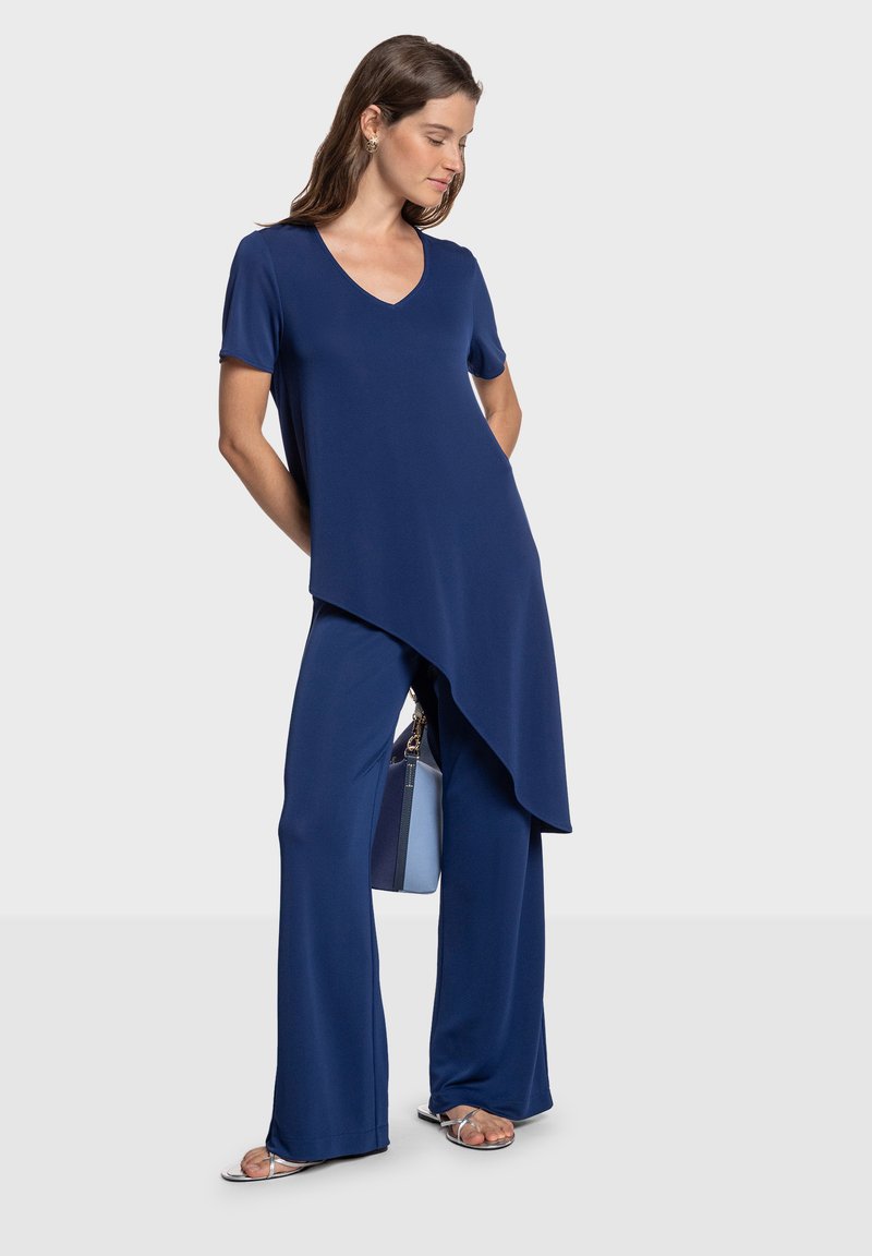 Tunique légère bleu marine avec encolure en V, manches courtes et ourlet asymétrique, associée à un pantalon à jambes larges de la même couleur. Sandales argentées et sac à main bleu.