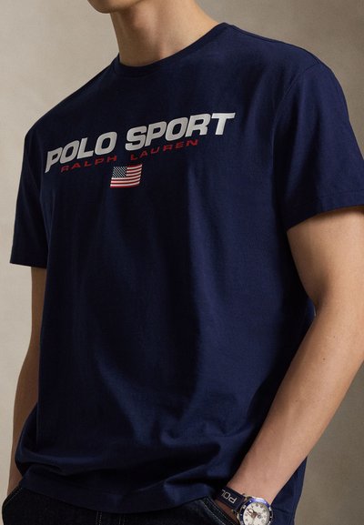 Μπλουζάκι από βαμβάκι σε ναυτικό μπλε χρώμα με τολμηρή επιγραφή "POLO SPORT" σε λευκά και κόκκινα γράμματα, με λεπτομέρεια τη σημαία των Ηνωμένων Πολιτειών. Κοντά μανίκια και στρογγυλή λαιμόκοψη.