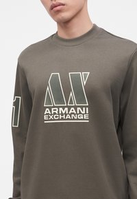 Νέος άνδρας που φοράει γκρι-πράσινο φούτερ Armani Exchange με μεγάλο γεωμετρικό λογότυπο και κείμενο στο στήθος και τον αριθμό ένα στο μανίκι.