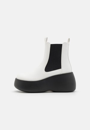 White ankle boots with a black elastic panel, chunky black platform sole, smooth texture, and rear pull tab for easy wear.