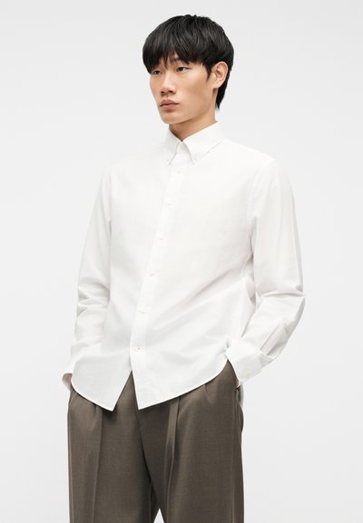 Νέος άνδρας που φοράει λευκό πουκάμισο με μακριά μανίκια και καφέ πλισέ παντελόνια, στέκεται με τα χέρια στις τσέπες μπροστά σε λευκό φόντο.