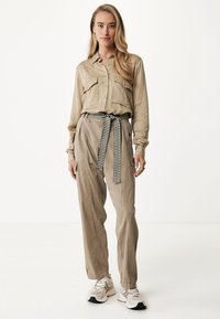 Beige košile na knoflíky s dvěma náprsními kapsami, v kombinaci se světle béžovými kalhotami a pruhovaným opaskem. Model má na sobě bílé tenisky.