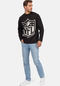 T-shirt nera a maniche lunghe con un grande logo NFL grigio sul davanti, indossata con jeans azzurri chiari e sneakers bianche.