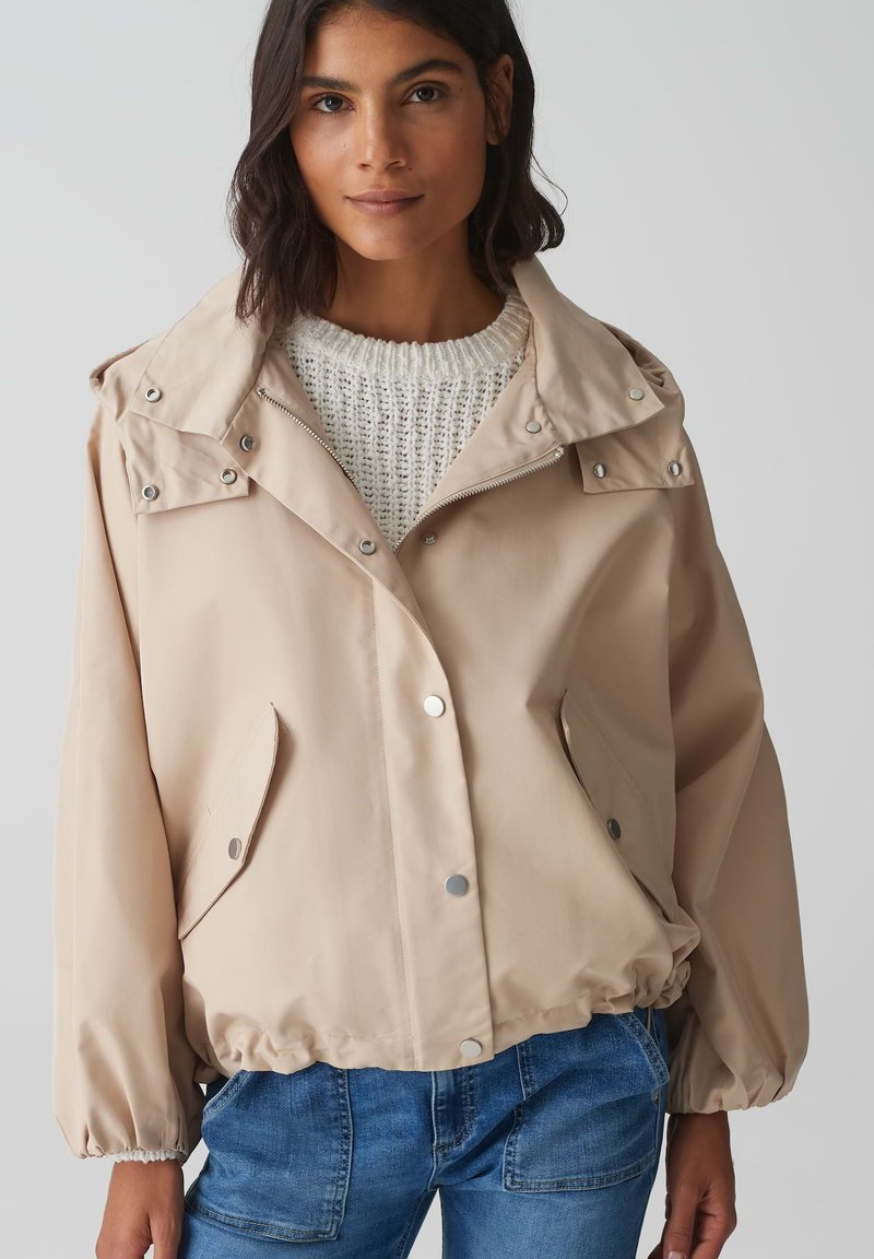 Giacca beige con cappuccio, bottoni a pressione e orlo elastico. Presenta due tasche laterali e un maglione di maglia bianca testurizzata sotto.
