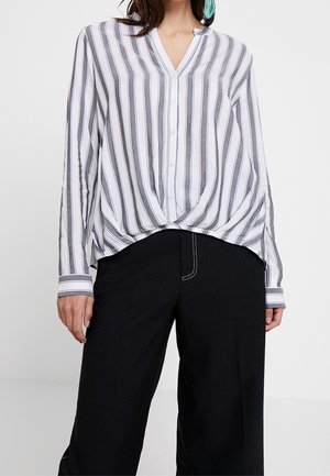 Femme portant un chemisier blanc boutonné avec des rayures grises verticales et un pantalon noir à jambes larges avec des coutures blanches visibles.