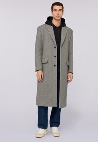 Szary wełniany płaszcz o strukturalnym designie, z dużymi klapami, dwiema kieszeniami i trzema guzikami, noszony na czarnej bluzie z kapturem, w zestawie z niebieskimi dżinsami i białymi sneakersami.