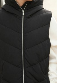 Czarna pikowana kamizelka z kapturem, z przeszywanym wzorem, zapinana na zamek z przodu i z bocznymi kieszeniami. Gładka tekstura i lekki materiał.