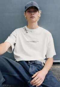 Tricou scurt de culoare gri deschis, cu detalii de stropi de cerneală neagră și text, purtat cu blugi negri și o șapcă gri, așezat lângă un perete.