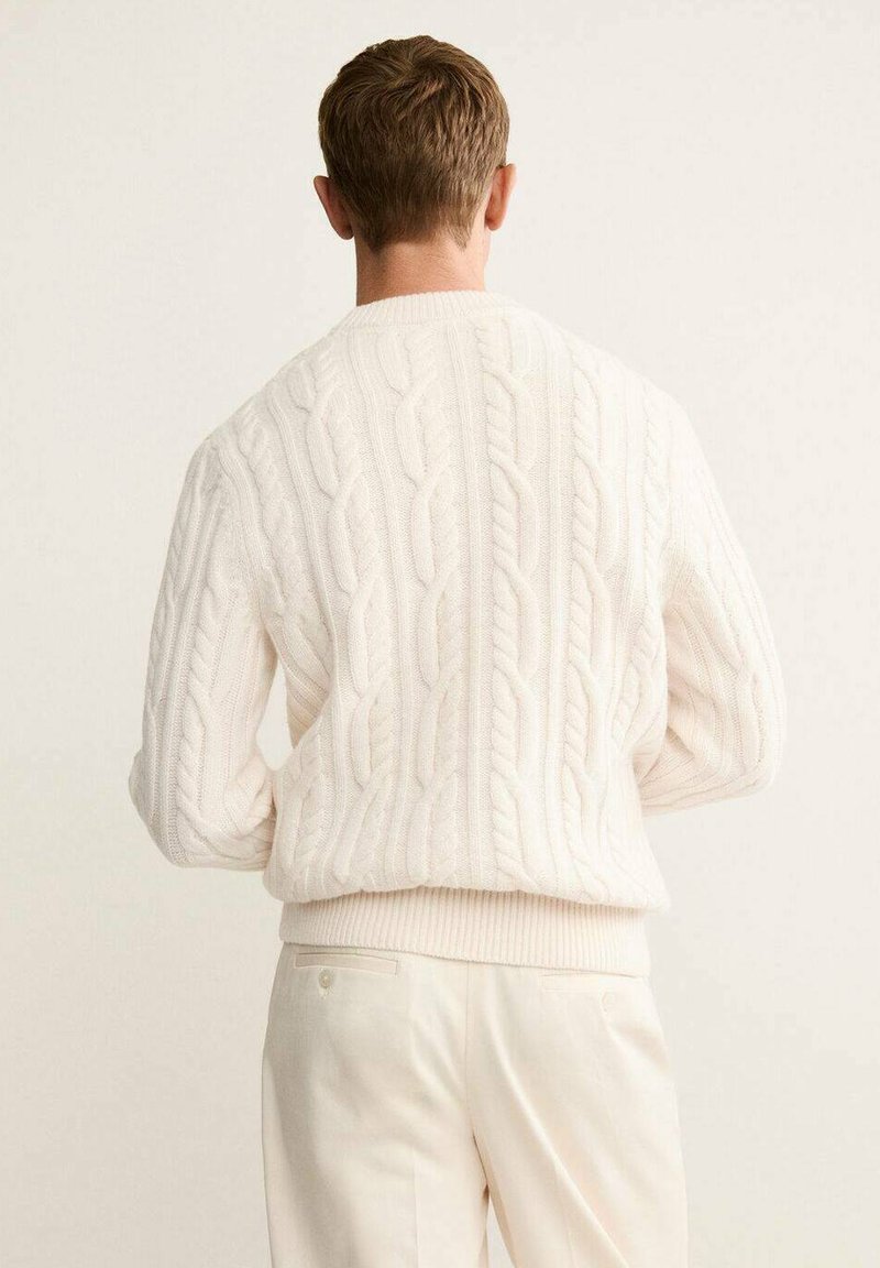 Cream cable-knit sweater with a ribbed hem and cuffs, featuring a textured pattern and a relaxed fit, worn with light-colored trousers.