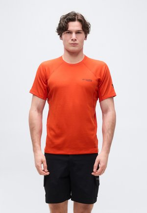 Jeune homme debout portant un t-shirt orange à manches courtes Columbia Titanium et un short noir sur un fond blanc uni.