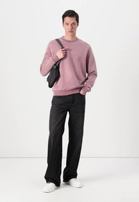 Helles pinkes Sweatshirt mit Rundhalsausschnitt und kleinem Logo, kombiniert mit lässigen schwarzen Jeans und weißen Sneakers; kleine schwarze Umhängetasche.