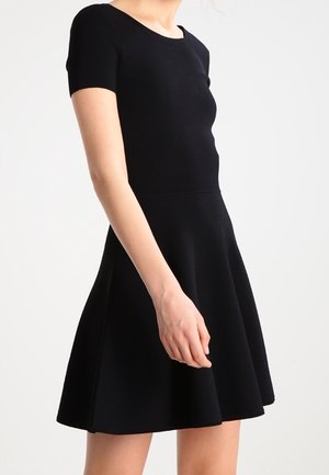 Femme portant une robe noire cintrée et évasée à manches courtes, debout, les bras détendus le long du corps, sur un fond uni.