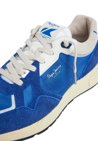 Modré tenisky s kombináciou semišu a nylonového materiálu. Obsahujú biele logo, krémové šnúrky a kontrastnú jemne bielu podrážku.