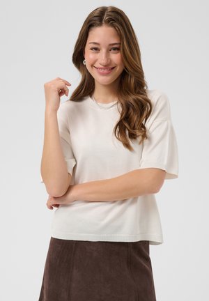 Jeune femme aux longs cheveux ondulés sourit, portant un top blanc à manches courtes, un collier argenté et une jupe marron sur un fond uni.