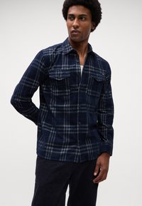 Man in een marineblauw en grijs geruit overhemd met twee borstzakken, donkere broek, die naar de zijkant kijkt tegen een effen achtergrond.