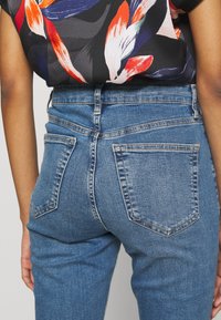 Vaqueros de mezclilla azul de talle alto con una textura suave, que presentan bolsillos traseros y detalles de costuras claras.