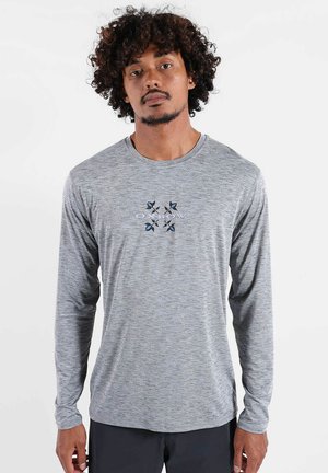 Homme aux cheveux bouclés portant un t-shirt gris à manches longues avec un petit motif floral bleu et noir et un texte sur la poitrine.