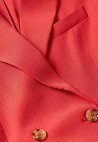 Blazer rouge sur mesure avec col cranté, deux boutons marron et une poche poitrine passepoilée, montrant la texture du tissu et les détails des coutures.