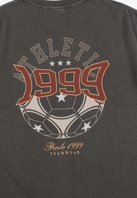 Ciemnoszary t-shirt z dużym graficznym wzorem zawierającym napis "ATHLETE 1999", piłkę nożną oraz "Prosto 1999 TEAMWEAR" w kolorze pomarańczowym i kremowym.