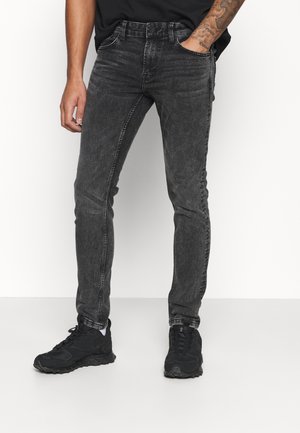 Homme portant un jean slim noir délavé et des baskets noires, debout avec une main dans la poche, sur fond blanc.