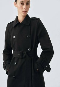 Trench coat negro de corte doble, con cinturón, que cuenta con grandes botones, hombreras y puños ajustables. Textura de tela suave.