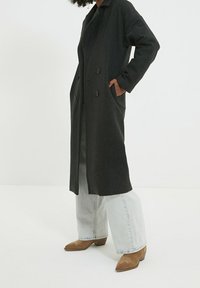 Lang mørkegrå frakke med dobbeltradet design, sidelommer og krave, båret over lysegrå brede jeans.