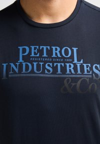 Námorně modré tričko vyrobené z měkké tkaniny, které má velký grafický potisk s textem "PETROL INDUSTRIES" v různých odstínech modré a černé.