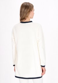Tunică crem cu mânecă lungă, confecționată dintr-un material moale, cu detalii bleu marin la decolteu și margini. Design simplu, croială dreaptă. Fără modele.