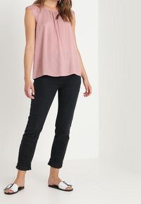 Růžový top bez rukávů s krajkovými detaily na ramenech, kombinovaný s černými zkrácenými kalhotami a bílými sandály s černými pásky.