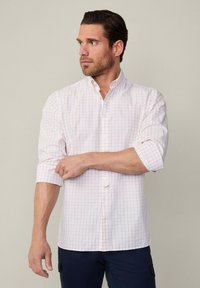 Camicia bianca button-up con una leggera fantasia a quadri rosa, maniche lunghe arrotolate fino ai gomiti e colletto classico. Indossata con pantaloni scuri.