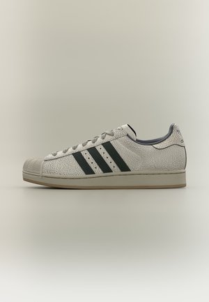 Baskets blanches Adidas avec des bandes noires, texture craquelée, bout renforcé en caoutchouc et semelle beige, présentées de profil sur fond neutre.