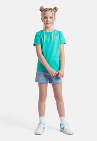 Νεαρή κοπέλα με ξανθά μαλλιά σε κότσους, φοράει τουρκουάζ μπλουζάκι Puma, τζιν σορτς, λευκές κάλτσες και λευκά αθλητικά παπούτσια με τουρκουάζ λεπτομέρειες.