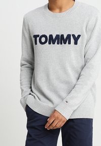 Maglione grigio chiaro con "TOMMY" in lettere navy in grassetto sul petto, indossato da una persona con le mani vicino alla vita, abbinato a pantaloni blu scuro.