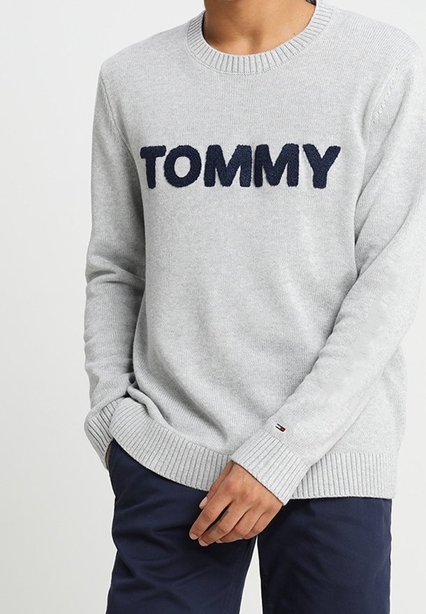Maglione grigio chiaro con "TOMMY" in lettere navy in grassetto sul petto, indossato da una persona con le mani vicino alla vita, abbinato a pantaloni blu scuro.