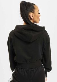 Sudadera corta negra con capucha, puños acanalados y dobladillo; presenta una textura suave y un corte relajado. Se lleva con pantalones negros y acentos blancos.