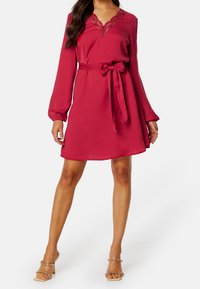 Robe rouge à manches longues, décolleté en dentelle et ceinture à la taille. Texture douce et coupe évasée. Portée avec des talons ouverts à bout doré.