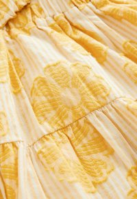 Tissu à motif floral jaune avec des fleurs en relief texturées sur un fond rayé clair, présentant des coutures froncées et un drapé fluide.