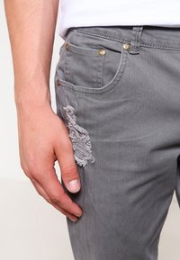 Šedé džíny z denim látky s vybledlým kusem látky na přední části. Obsahují zlaté kovové doplňky a klasický design s pěti kapsami.