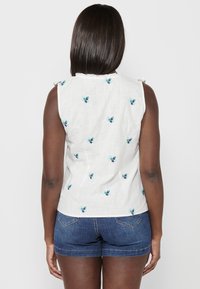Weißes ärmelloses Oberteil mit floralen Stickereien in Blau und Türkis, mit einer strukturierten Oberfläche und Rüschendetails an den Schultern. Kombiniert mit Jeansshorts.