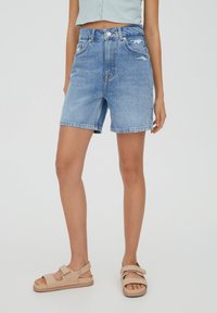 Shorts de mezclilla azul claro con cintura alta, detalles desgastados y dobladillo deshilachado. Combinados con sandalias beige y un top corto verde claro.