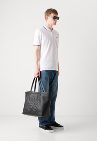 Borsa tote in pelle nera con logo "HUGO" in rilievo, forma piatta, doppi manici e una texture liscia. Il modello indossa una polo bianca e jeans blu.