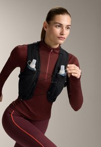 Čierna hydratačná vesta s dvoma vreckami na fľaše, nastaviteľnými popruhmi a aerodynamickým dizajnom, nosená cez bordový, textúrovaný dlhý rukáv.