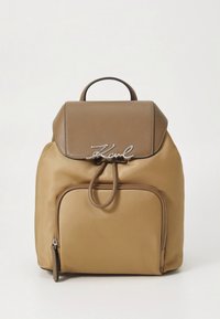 Beige nylonryggsäck med brun konstläderklaff, silverdetaljer, framficka med dragkedja och snörning i toppen.