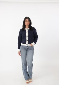 Cardigan lavorato a maglia blu navy con tessuto a trama, chiusure con bottoni bianchi e due tasche frontali, abbinato a jeans azzurri e sandali bianchi.